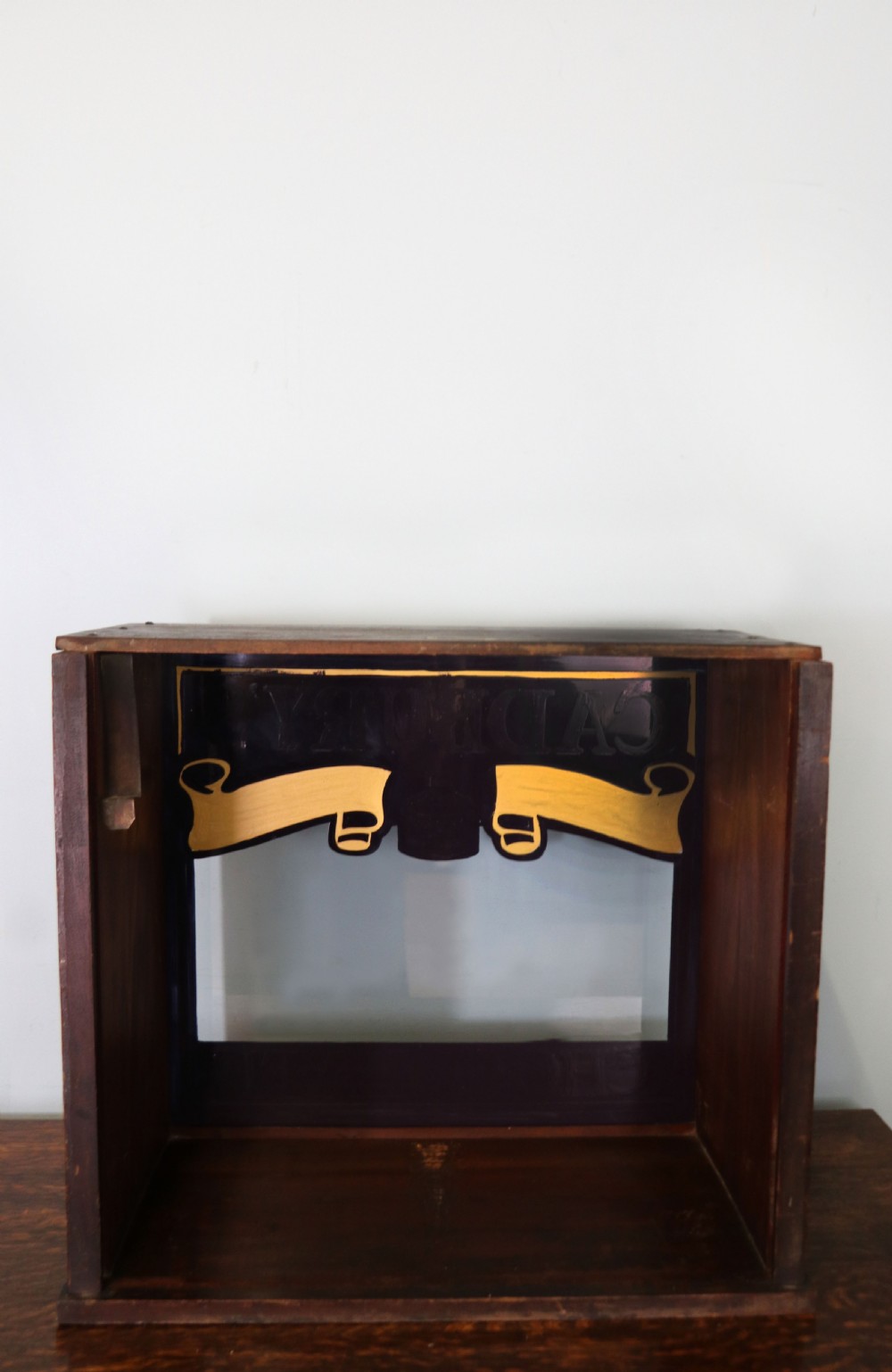 A dark hardwood rectangular shop display cabinet featuring gold lettering on the front glass pane reading Cadburys Dairy Milk Chocolate. View VI