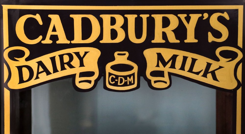 A dark hardwood rectangular shop display cabinet featuring gold lettering on the front glass pane reading Cadburys Dairy Milk Chocolate. View VII