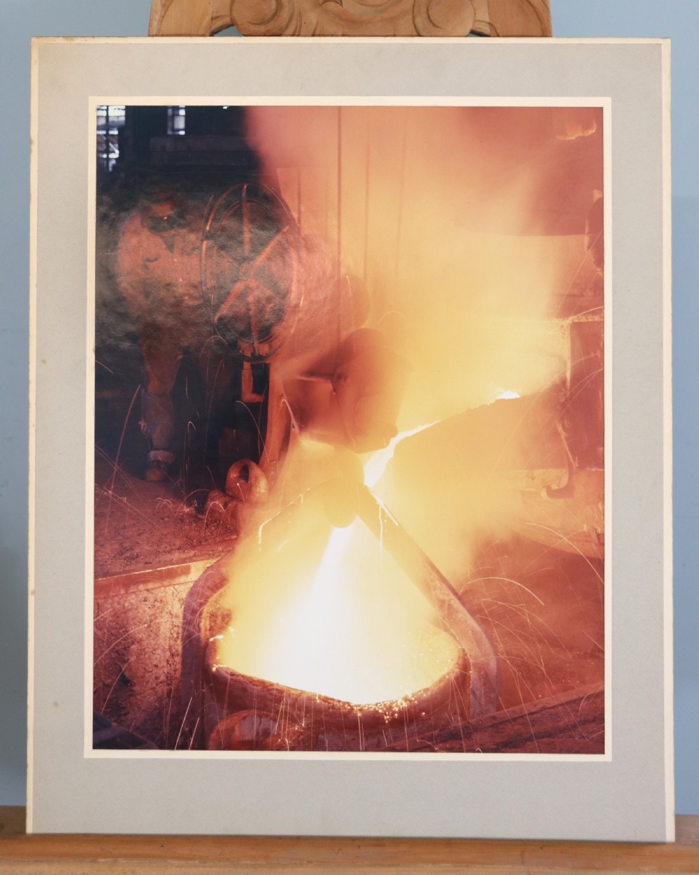 A mounted mid-century colour photograph depicting a foundry worker pouring molten metal. View II