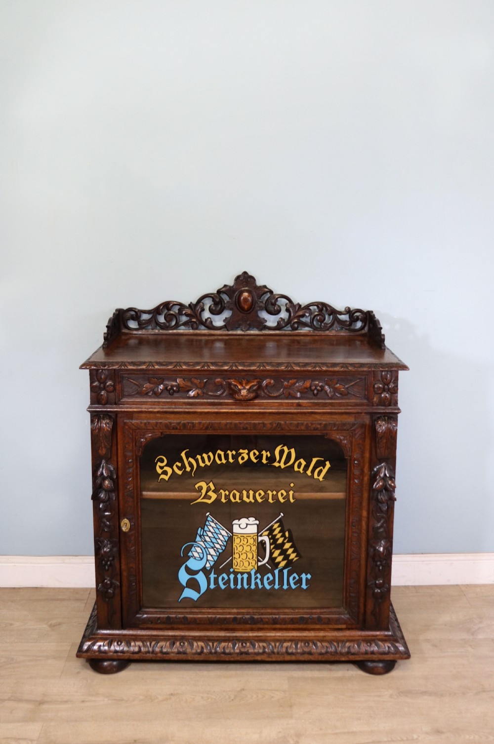 A dark carved oak side cabinet with a single glazed door displaying painted brewery signwriting and an ornate carved gallery back.