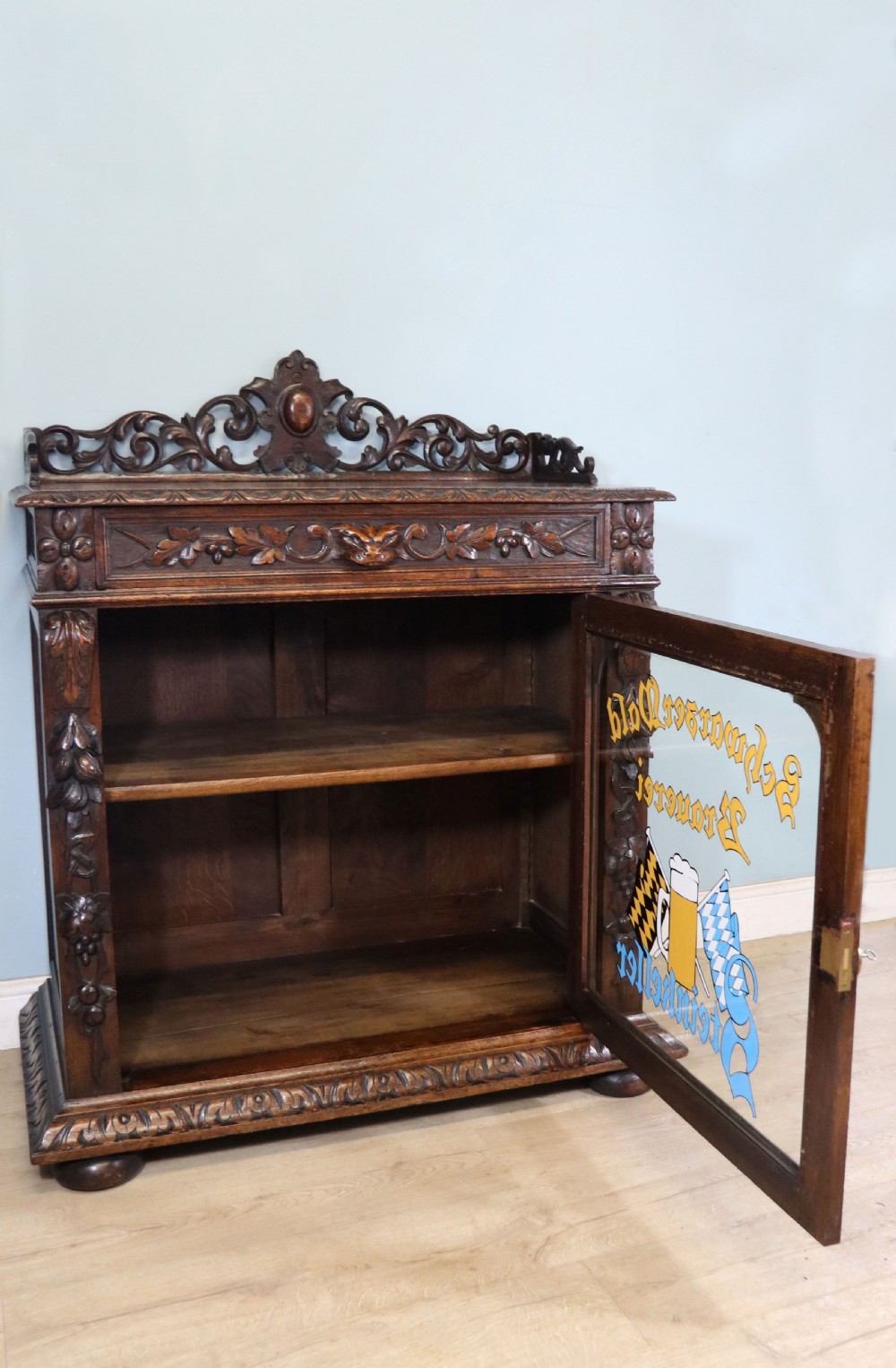 A dark carved oak side cabinet with a single glazed door displaying painted brewery signwriting and an ornate carved gallery back. View IV