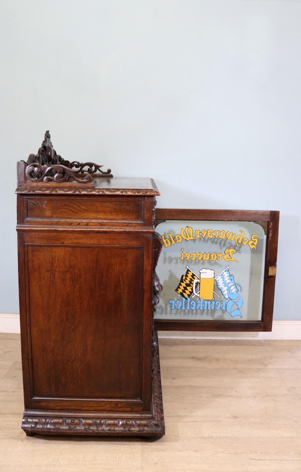 A dark carved oak side cabinet with a single glazed door displaying painted brewery signwriting and an ornate carved gallery back. View VI