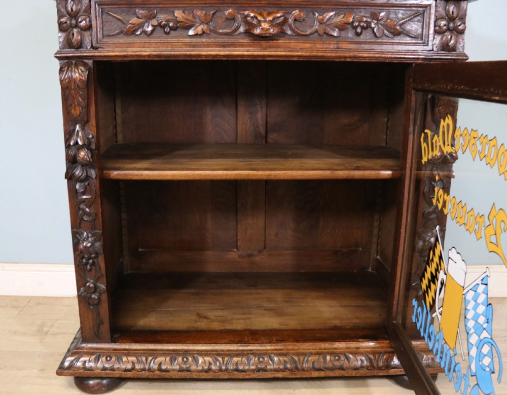 A dark carved oak side cabinet with a single glazed door displaying painted brewery signwriting and an ornate carved gallery back. View XXX