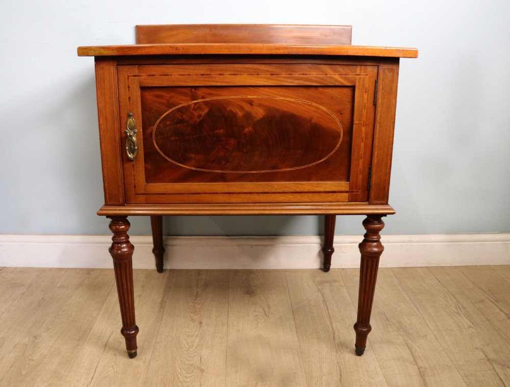 An Edwardian mahogany bedside cabinet with a single door containing a central oval marquetry inlay, raised on fluted turned legs. View II