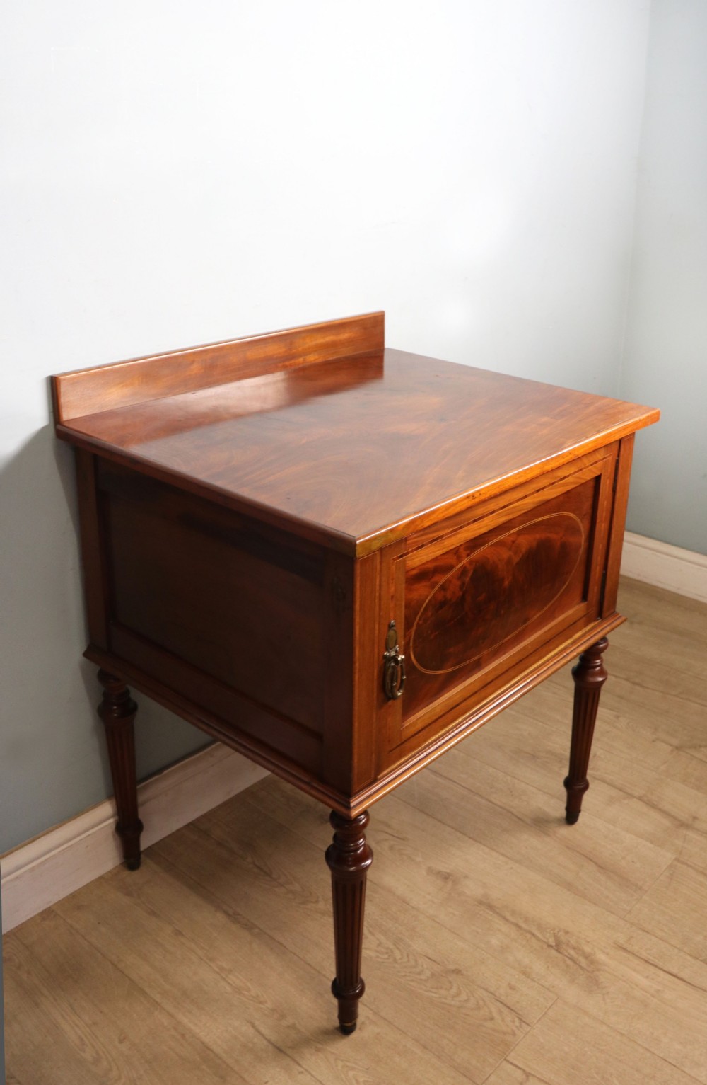 An Edwardian mahogany bedside cabinet with a single door containing a central oval marquetry inlay, raised on fluted turned legs. View VI