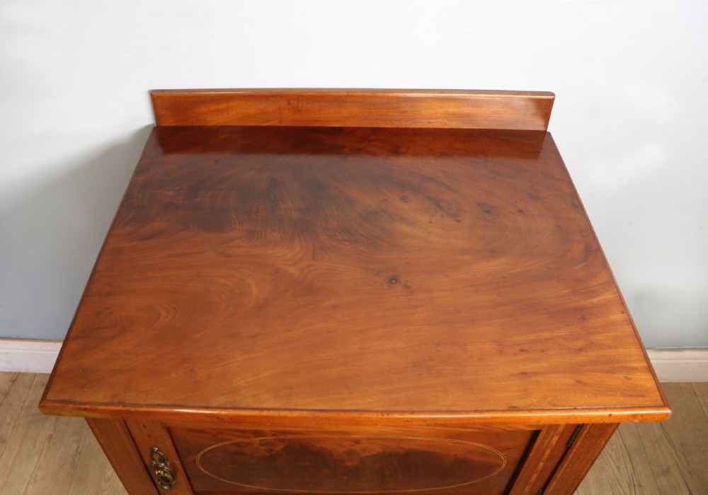 An Edwardian mahogany bedside cabinet with a single door containing a central oval marquetry inlay, raised on fluted turned legs. View VIII