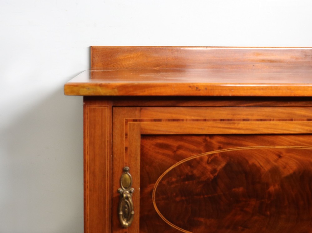 An Edwardian mahogany bedside cabinet with a single door containing a central oval marquetry inlay, raised on fluted turned legs. View XI