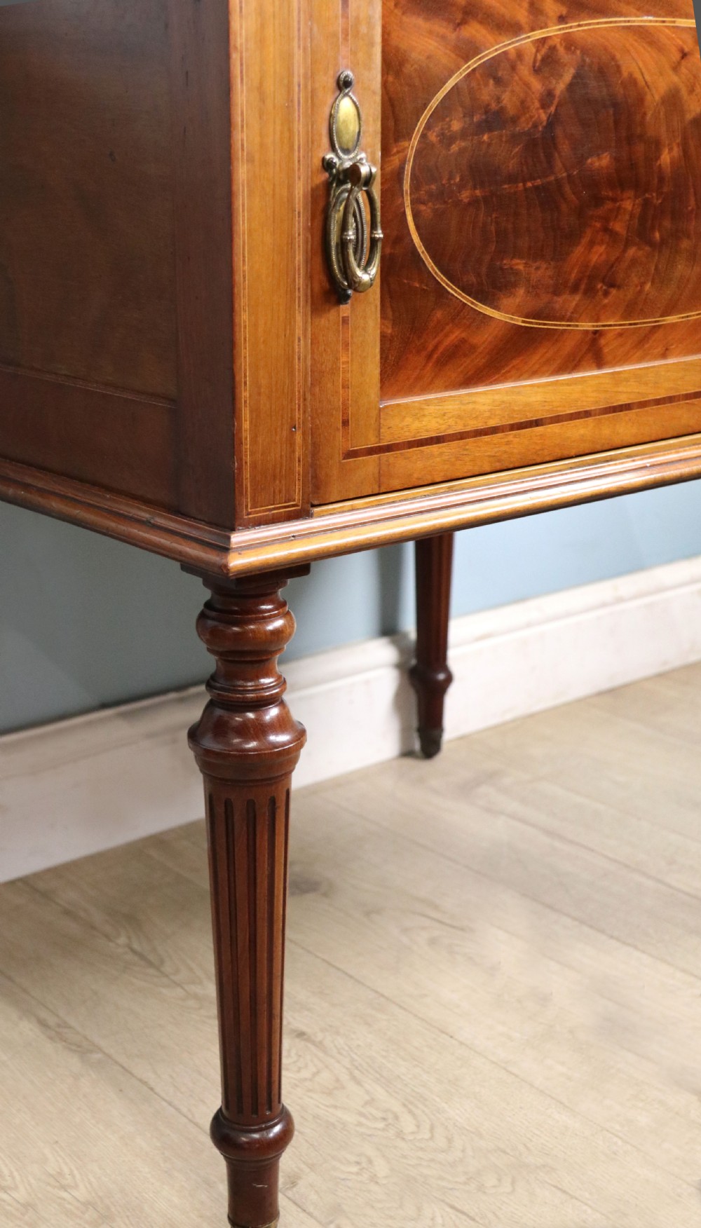 An Edwardian mahogany bedside cabinet with a single door containing a central oval marquetry inlay, raised on fluted turned legs. View XVI