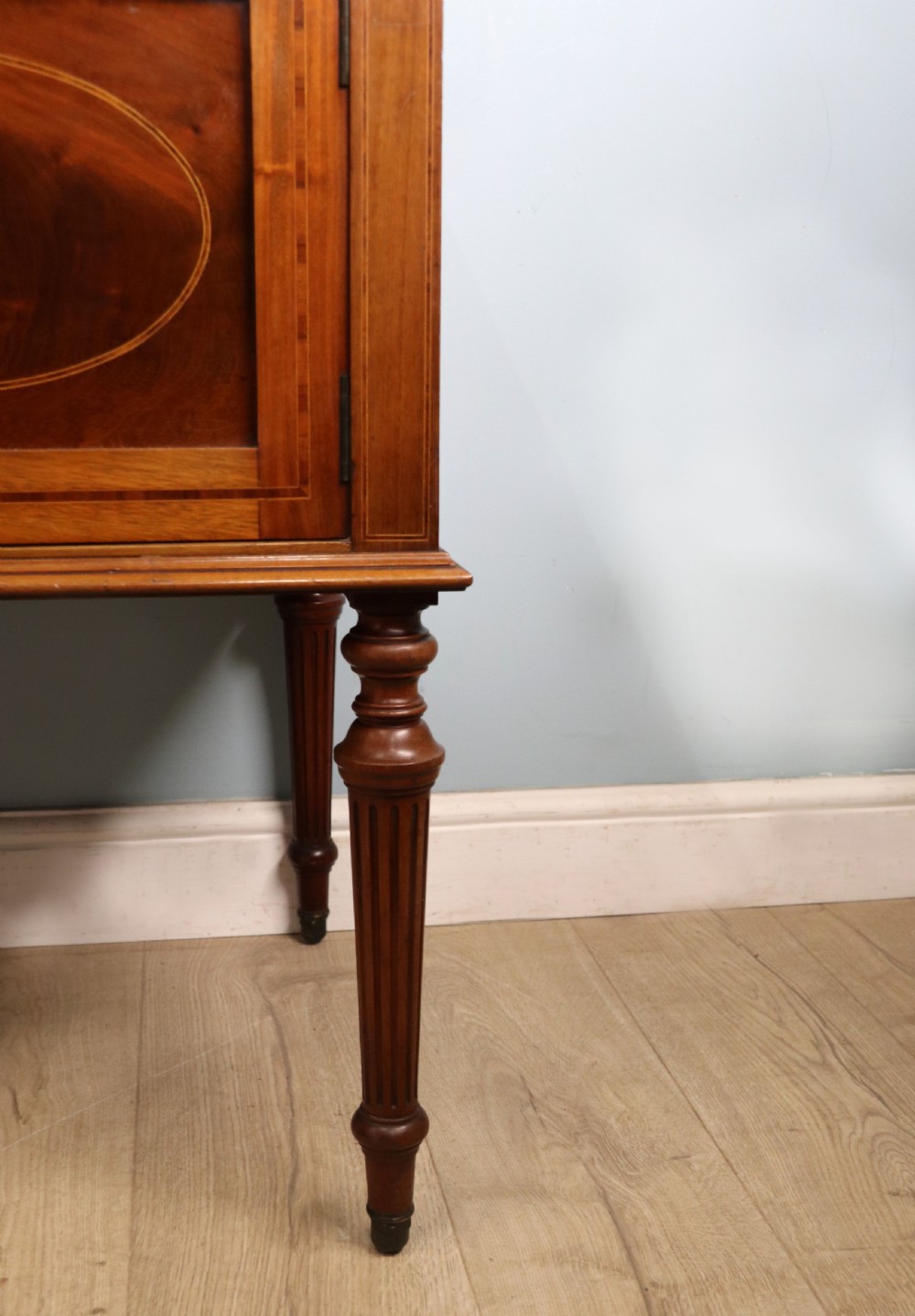An Edwardian mahogany bedside cabinet with a single door containing a central oval marquetry inlay, raised on fluted turned legs. View XVII