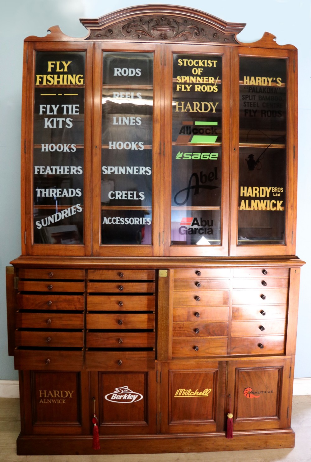A tall Victorian solid walnut shop cabinet with glazed upper doors featuring sign-written fishing typography, multiple small drawers in the midsection, and panelled cupboard doors below. View V