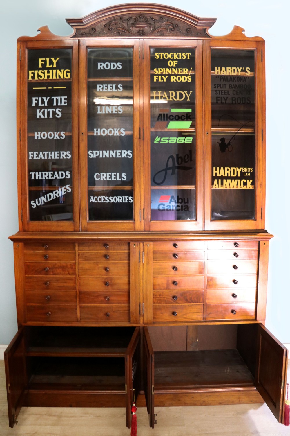 A tall Victorian solid walnut shop cabinet with glazed upper doors featuring sign-written fishing typography, multiple small drawers in the midsection, and panelled cupboard doors below. View VI