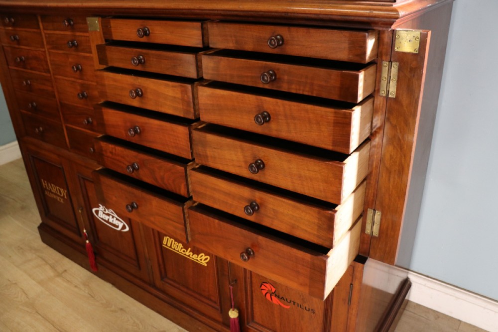 A tall Victorian solid walnut shop cabinet with glazed upper doors featuring sign-written fishing typography, multiple small drawers in the midsection, and panelled cupboard doors below. View XIII