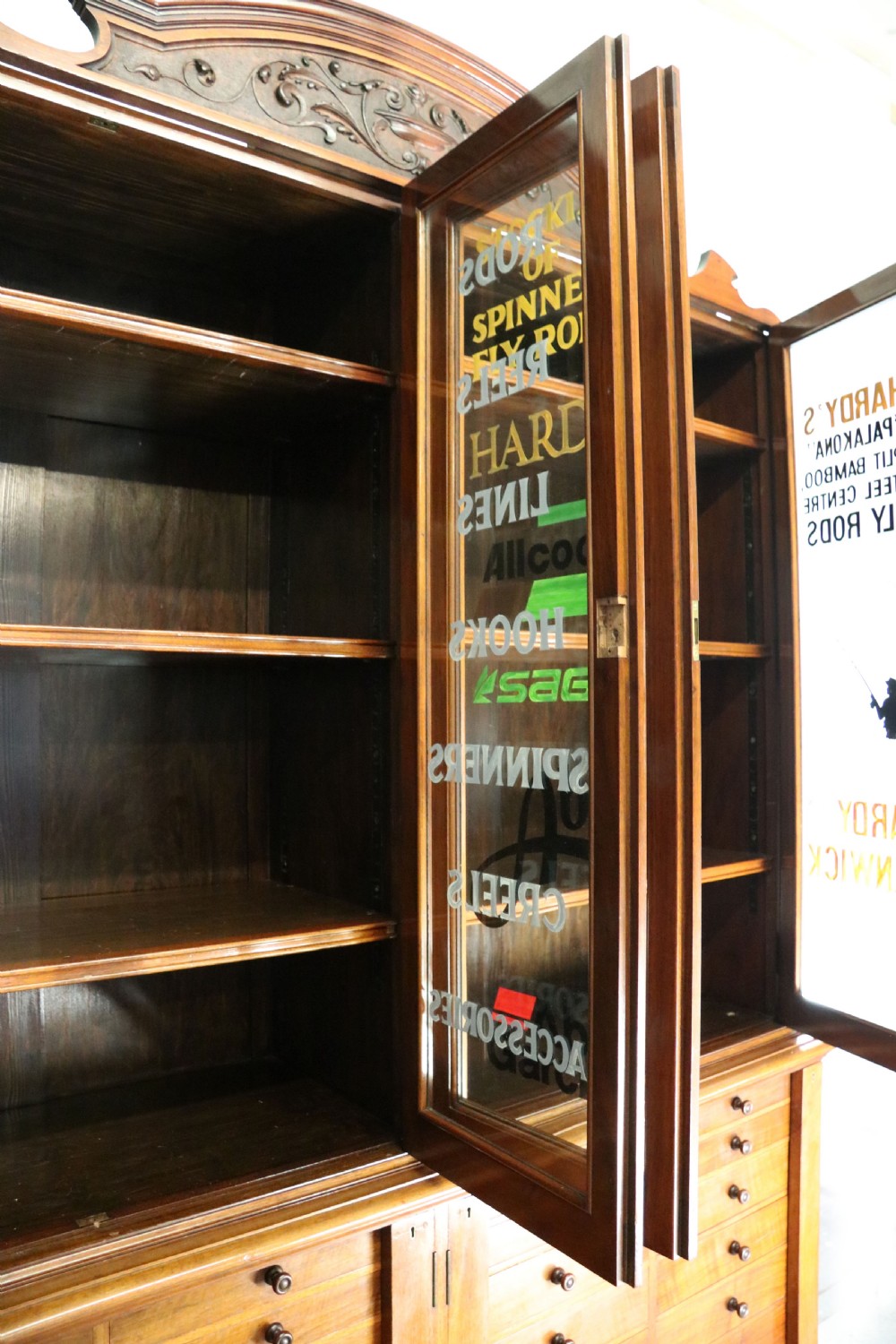 A tall Victorian solid walnut shop cabinet with glazed upper doors featuring sign-written fishing typography, multiple small drawers in the midsection, and panelled cupboard doors below. View XVII
