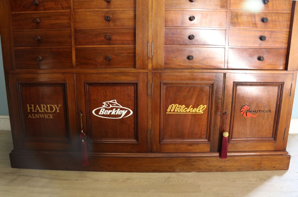 A tall Victorian solid walnut shop cabinet with glazed upper doors featuring sign-written fishing typography, multiple small drawers in the midsection, and panelled cupboard doors below. View XXVIII
