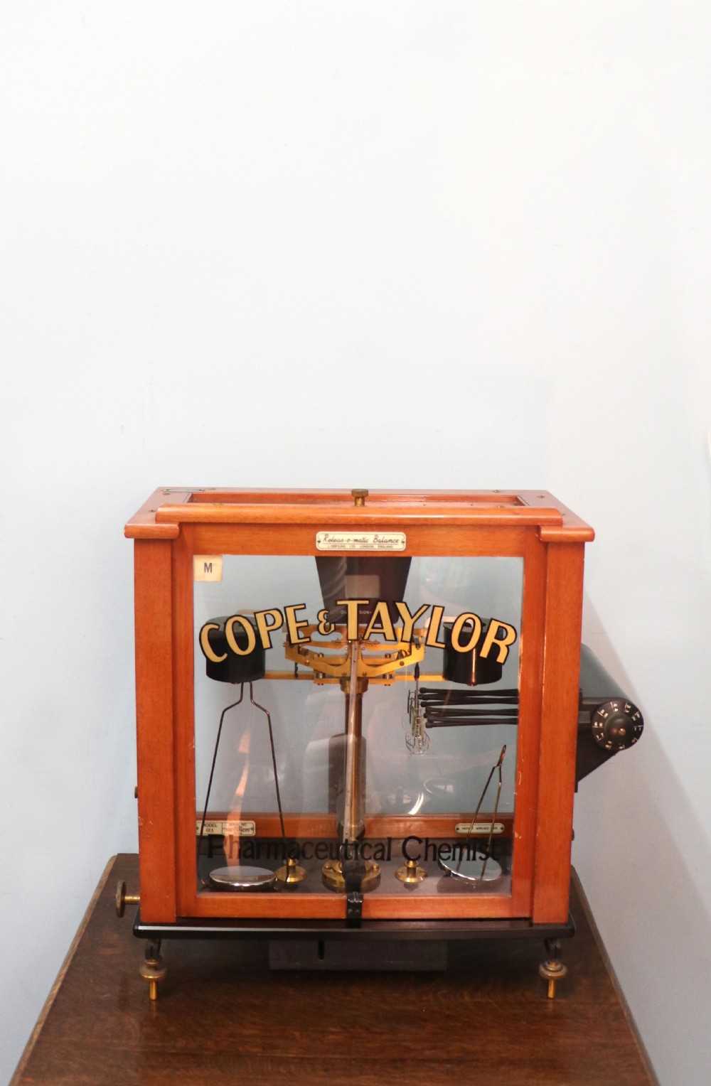 An Oertling precision chemists beam balance scale with a brass mechanism housed inside a glazed timber cabinet featuring gilded gold typography.
