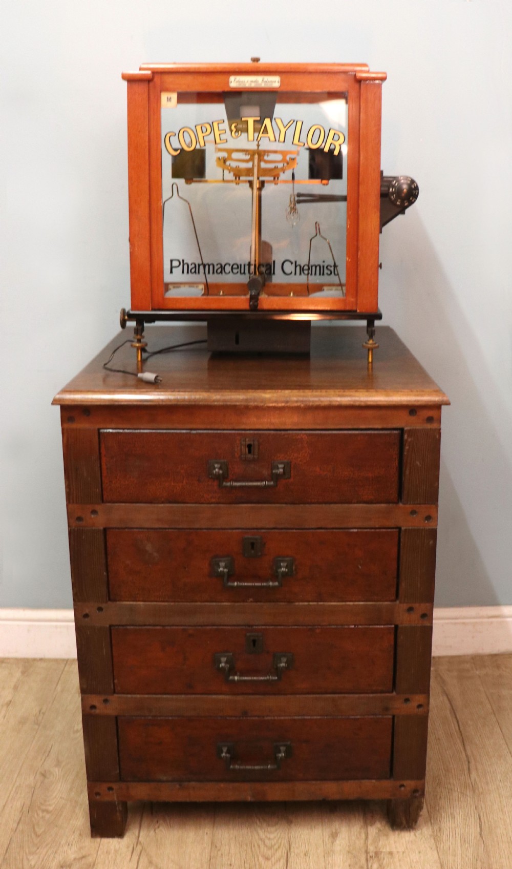 An Oertling precision chemists beam balance scale with a brass mechanism housed inside a glazed timber cabinet featuring gilded gold typography. View II