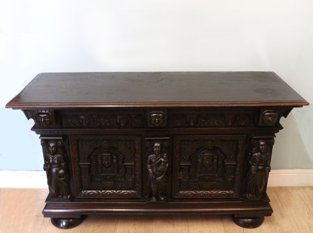 A dark carved Flemish oak side cabinet featuring detailed figural caryatids, relief mascarons, architectural door panels, and heavy bun feet.