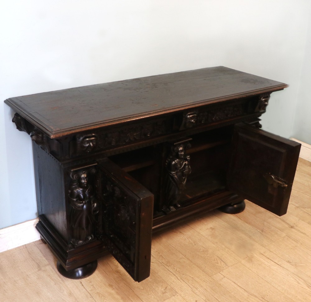 A dark carved Flemish oak side cabinet featuring detailed figural caryatids, relief mascarons, architectural door panels, and heavy bun feet. View IV