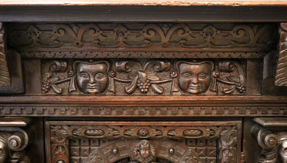 A dark carved Flemish oak side cabinet featuring detailed figural caryatids, relief mascarons, architectural door panels, and heavy bun feet. View IX