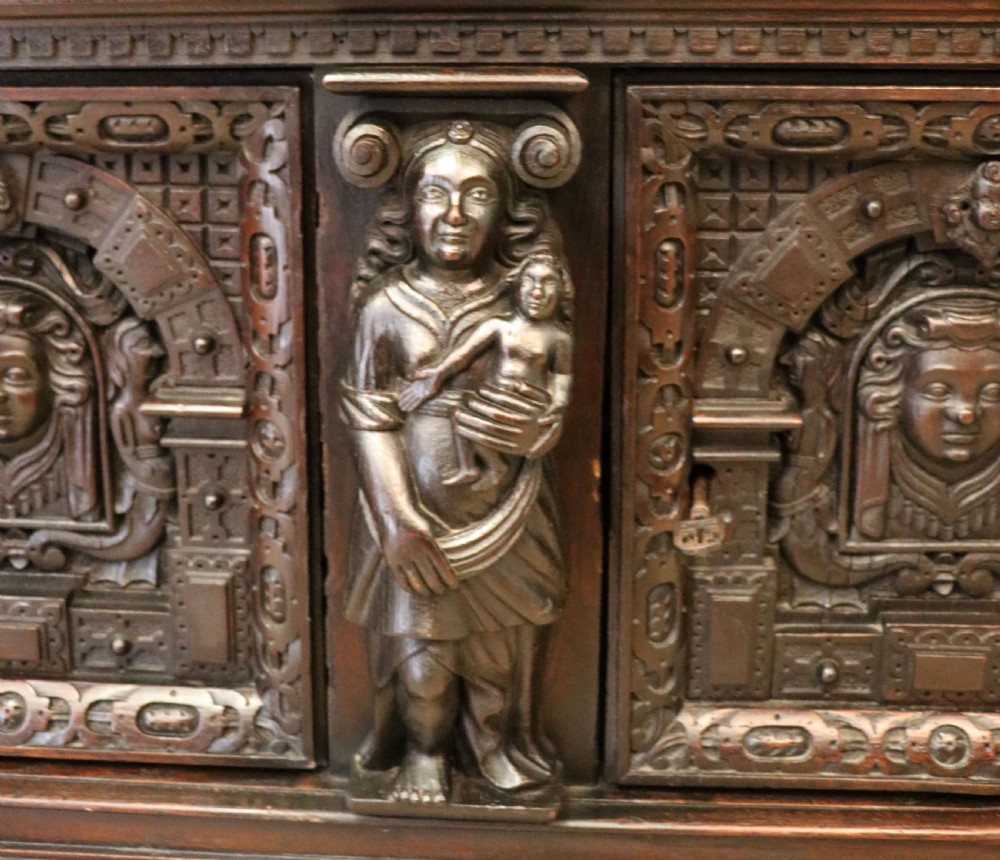 A dark carved Flemish oak side cabinet featuring detailed figural caryatids, relief mascarons, architectural door panels, and heavy bun feet. View XIII