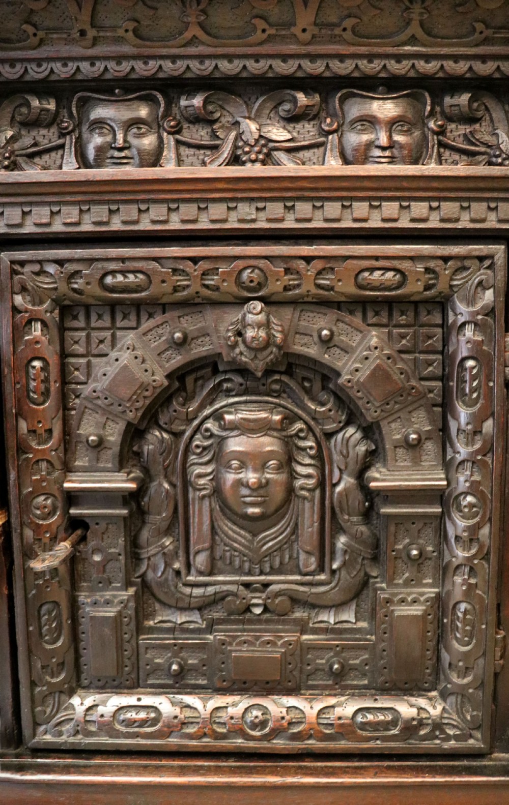 A dark carved Flemish oak side cabinet featuring detailed figural caryatids, relief mascarons, architectural door panels, and heavy bun feet. View XV