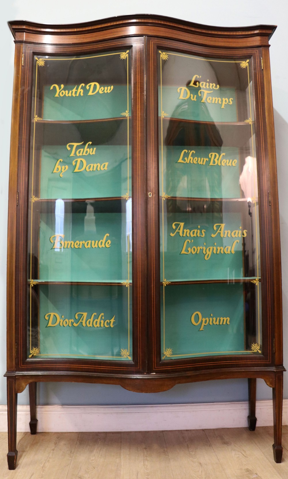 An Edwardian mahogany display cabinet featuring double glazed doors with gold sign-written perfume brand names against a turquoise painted interior. View II