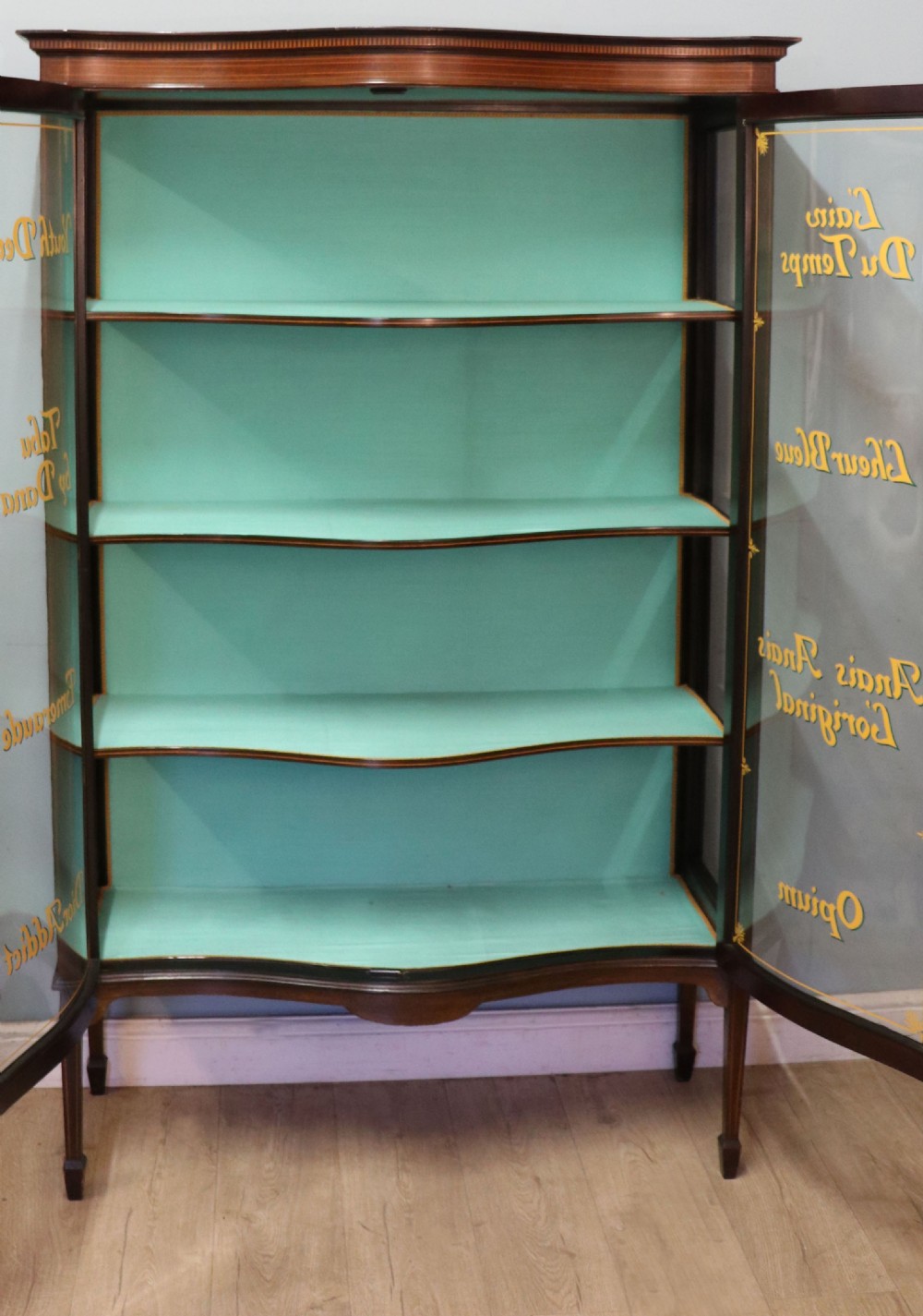 An Edwardian mahogany display cabinet featuring double glazed doors with gold sign-written perfume brand names against a turquoise painted interior. View IV