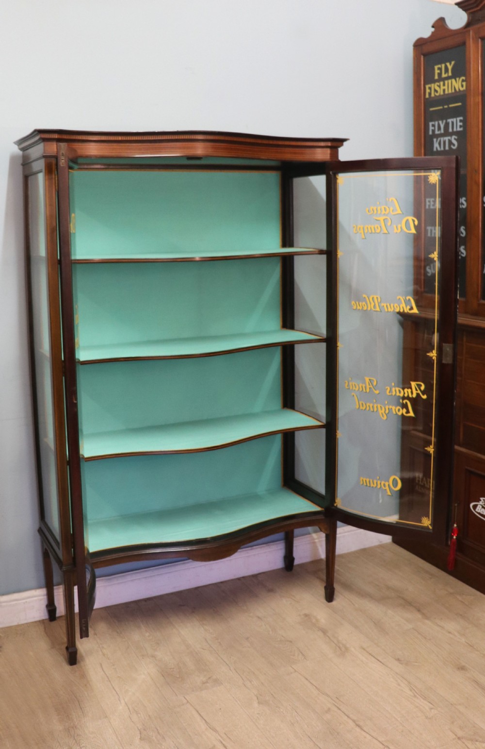 An Edwardian mahogany display cabinet featuring double glazed doors with gold sign-written perfume brand names against a turquoise painted interior. View V