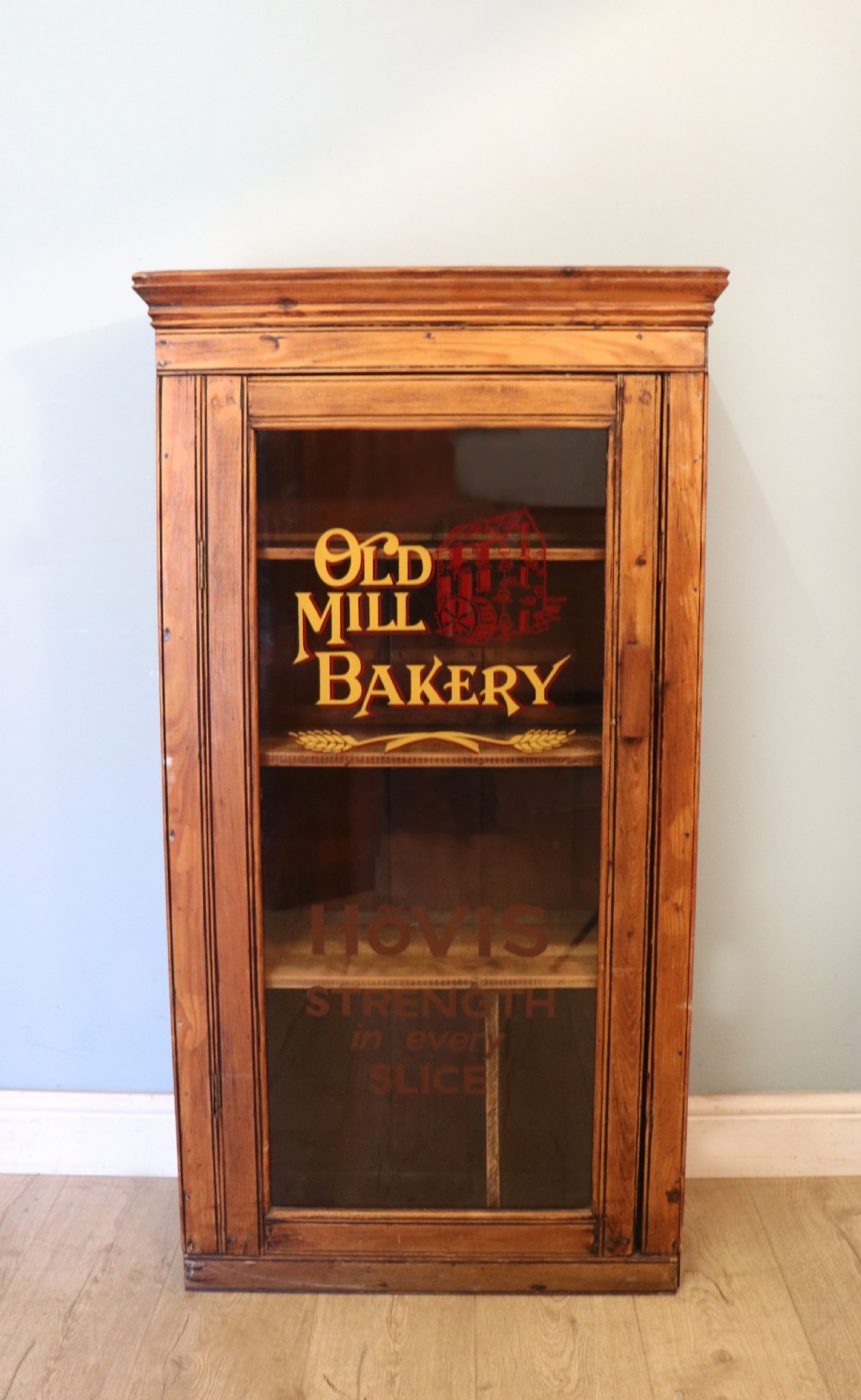 A glazed pine cabinet featuring a glass door with gold and red bakery signwriting and internal wooden shelves.