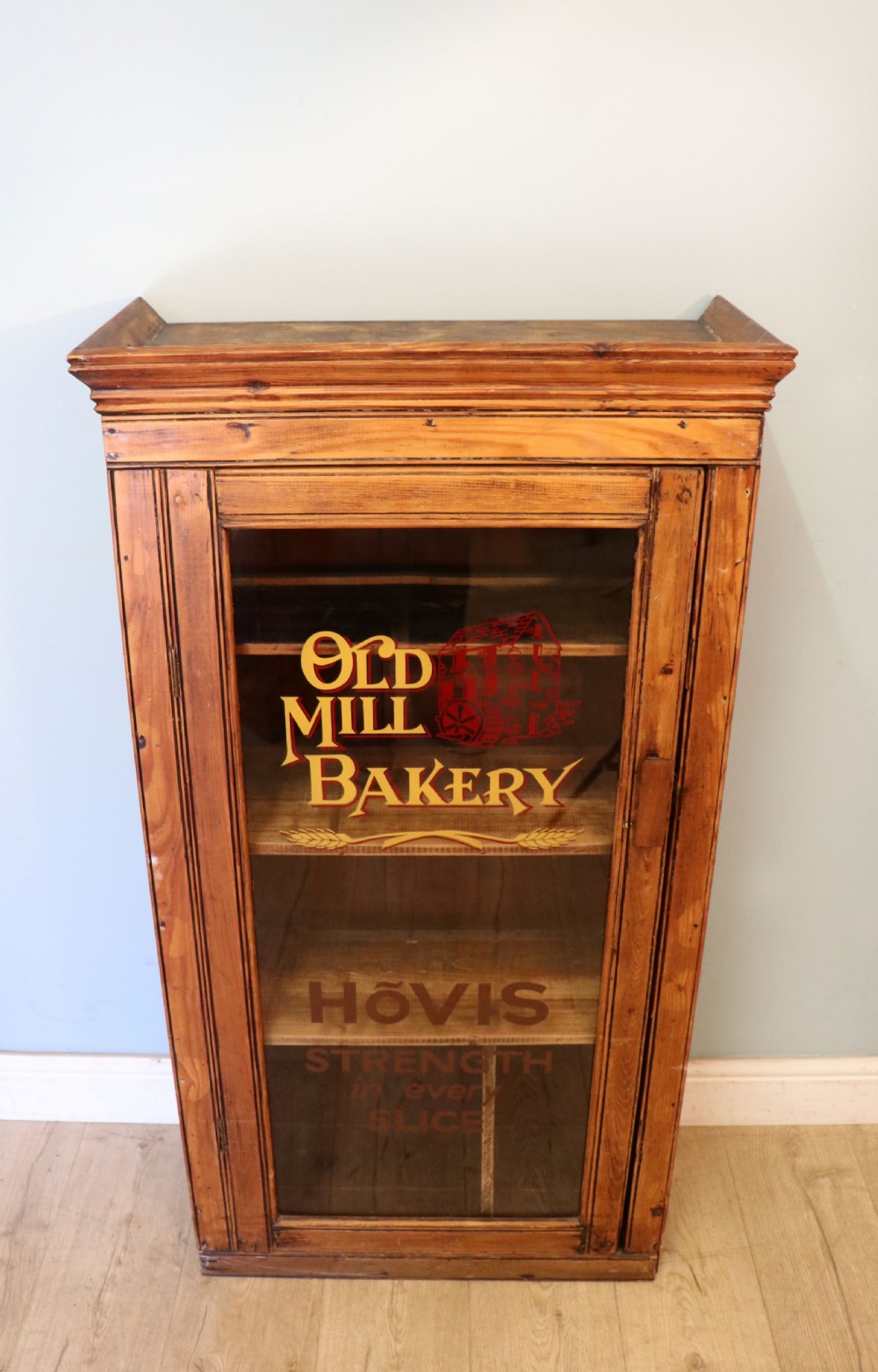 A glazed pine cabinet featuring a glass door with gold and red bakery signwriting and internal wooden shelves. View II
