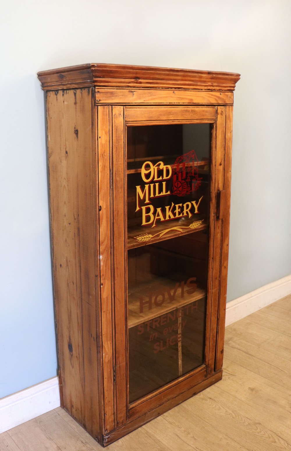 A glazed pine cabinet featuring a glass door with gold and red bakery signwriting and internal wooden shelves. View III