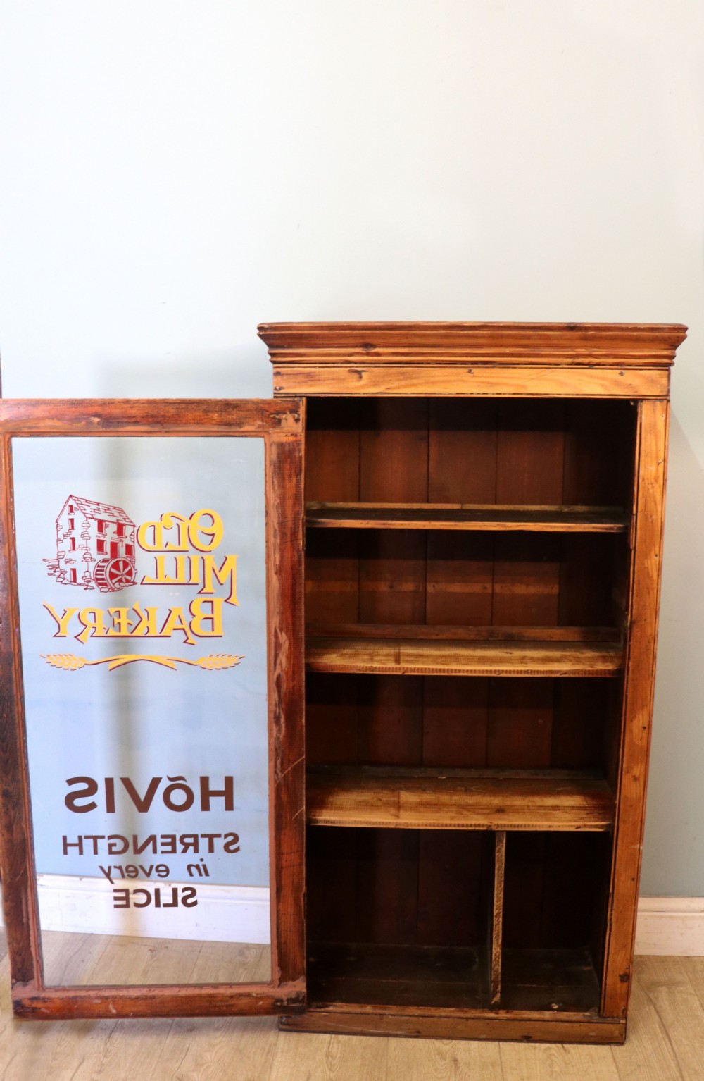 A glazed pine cabinet featuring a glass door with gold and red bakery signwriting and internal wooden shelves. View V