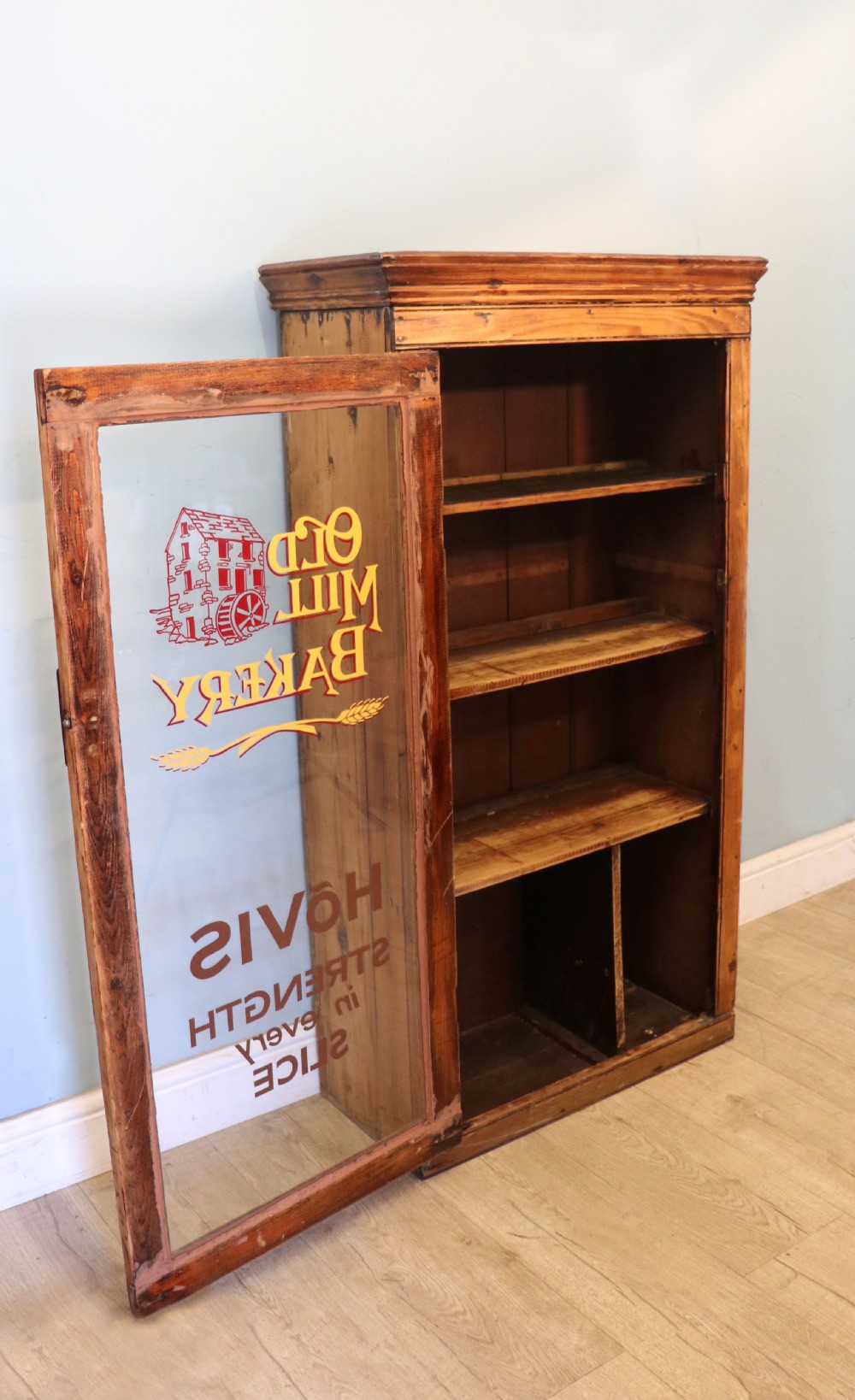A glazed pine cabinet featuring a glass door with gold and red bakery signwriting and internal wooden shelves. View VI