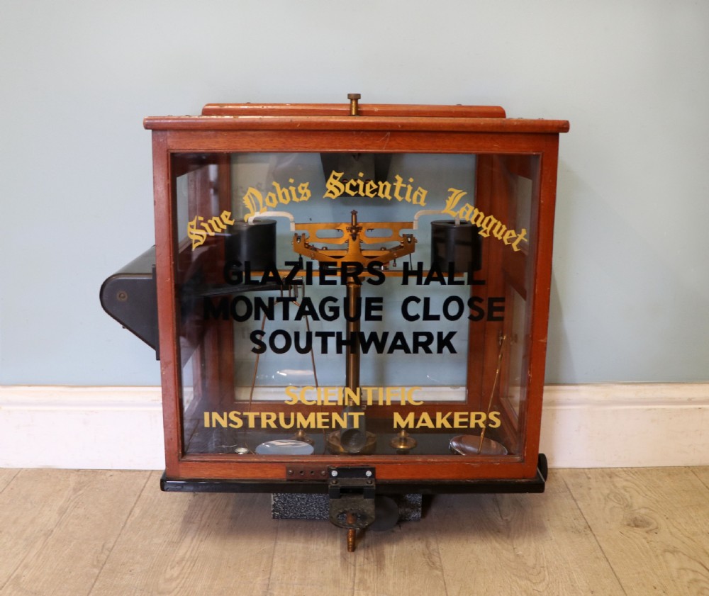 A set of brass chemist precision beam scales enclosed within a glazed mahogany display case featuring gilt and black sign-written lettering.