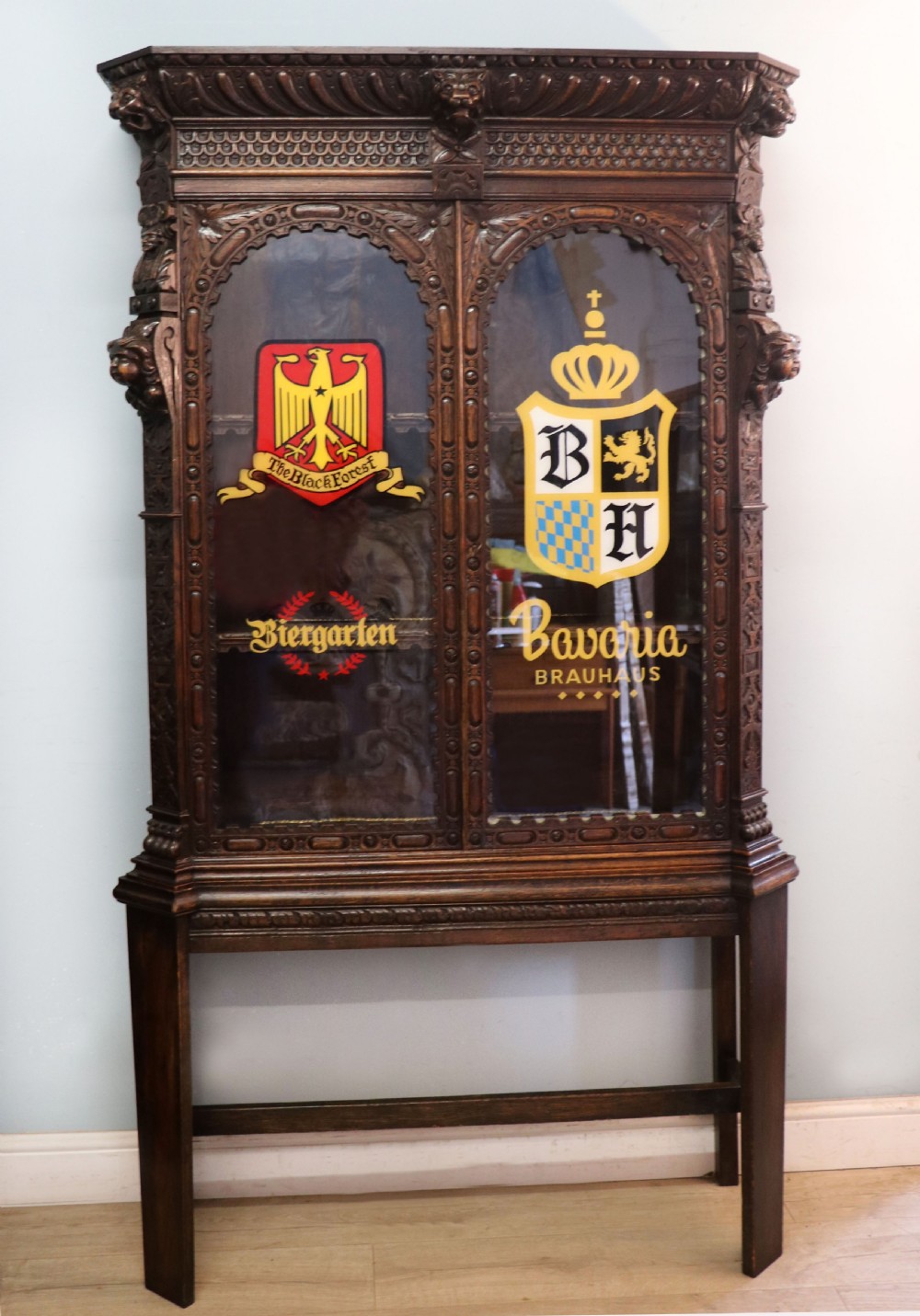 A late 19th-century dark oak glazed display cabinet on a tall stand featuring detailed lion mask relief carvings and hand-painted German heraldic emblems on the glass doors.