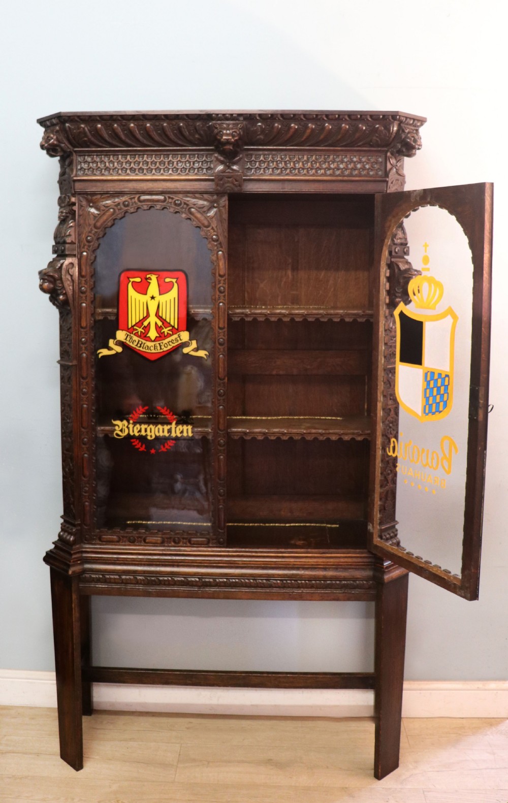 A late 19th-century dark oak glazed display cabinet on a tall stand featuring detailed lion mask relief carvings and hand-painted German heraldic emblems on the glass doors. View II