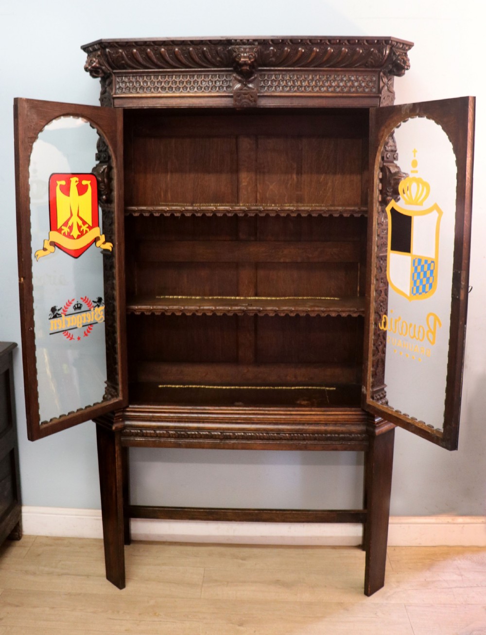 A late 19th-century dark oak glazed display cabinet on a tall stand featuring detailed lion mask relief carvings and hand-painted German heraldic emblems on the glass doors. View III