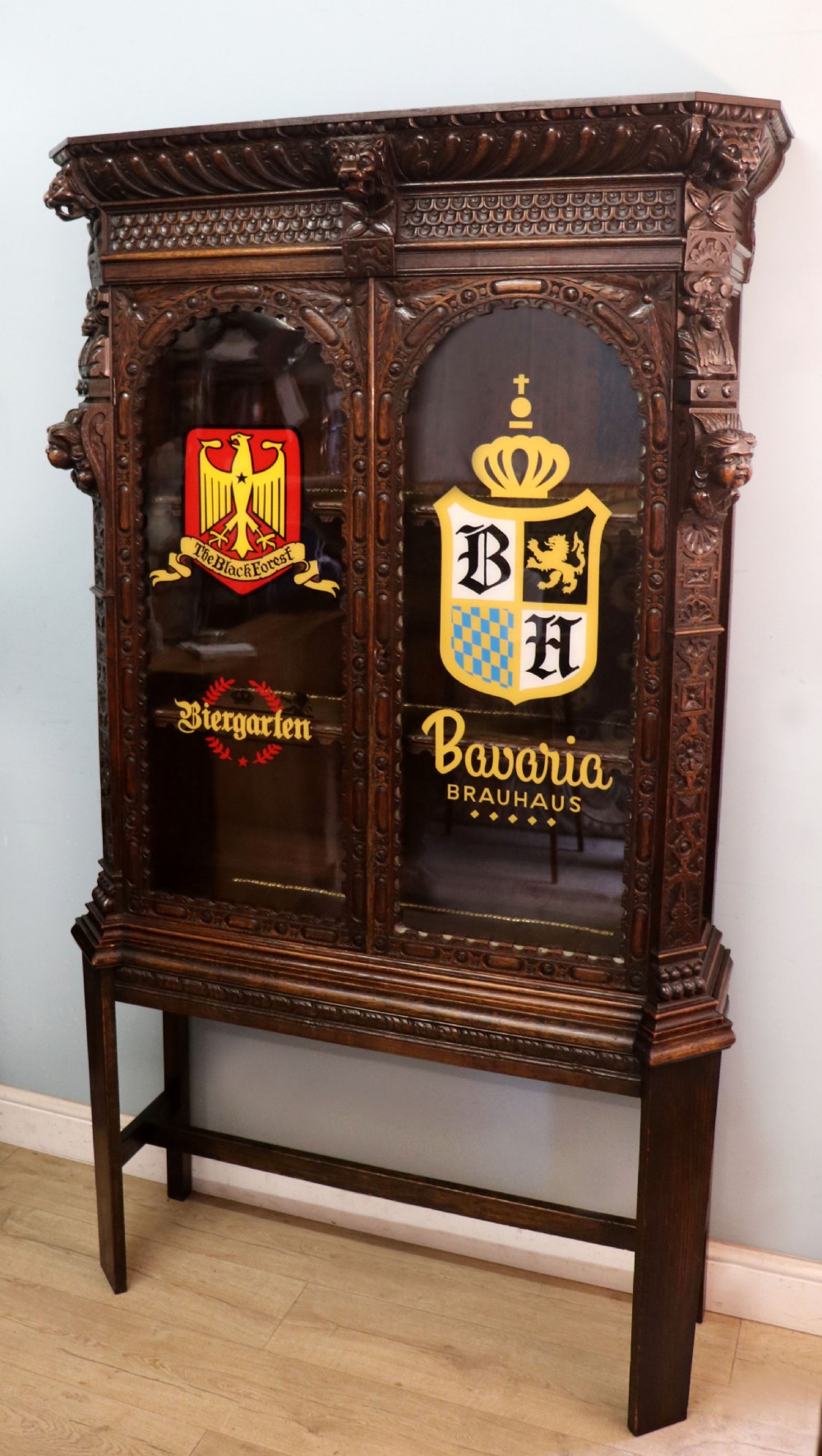 A late 19th-century dark oak glazed display cabinet on a tall stand featuring detailed lion mask relief carvings and hand-painted German heraldic emblems on the glass doors. View VI