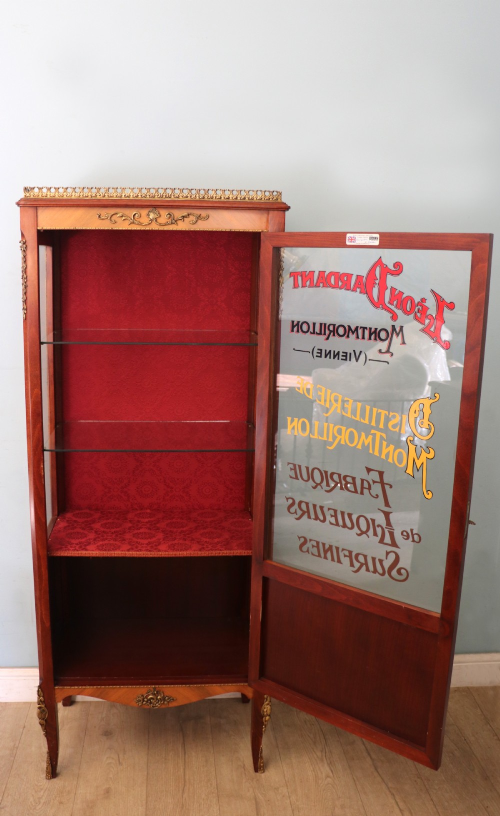 A French Vernis Martin style mahogany vitrine cabinet featuring a brass top gallery, glass shelves, red fabric lining, and a glazed door with gilded lettering.