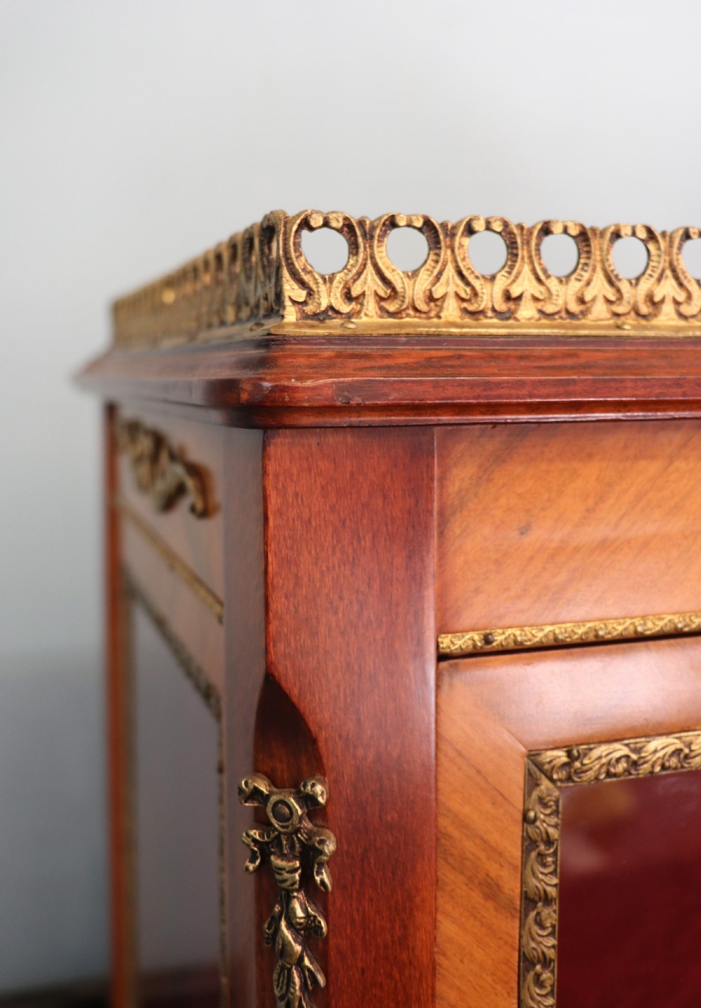 A French Vernis Martin style mahogany vitrine cabinet featuring a brass top gallery, glass shelves, red fabric lining, and a glazed door with gilded lettering. View V