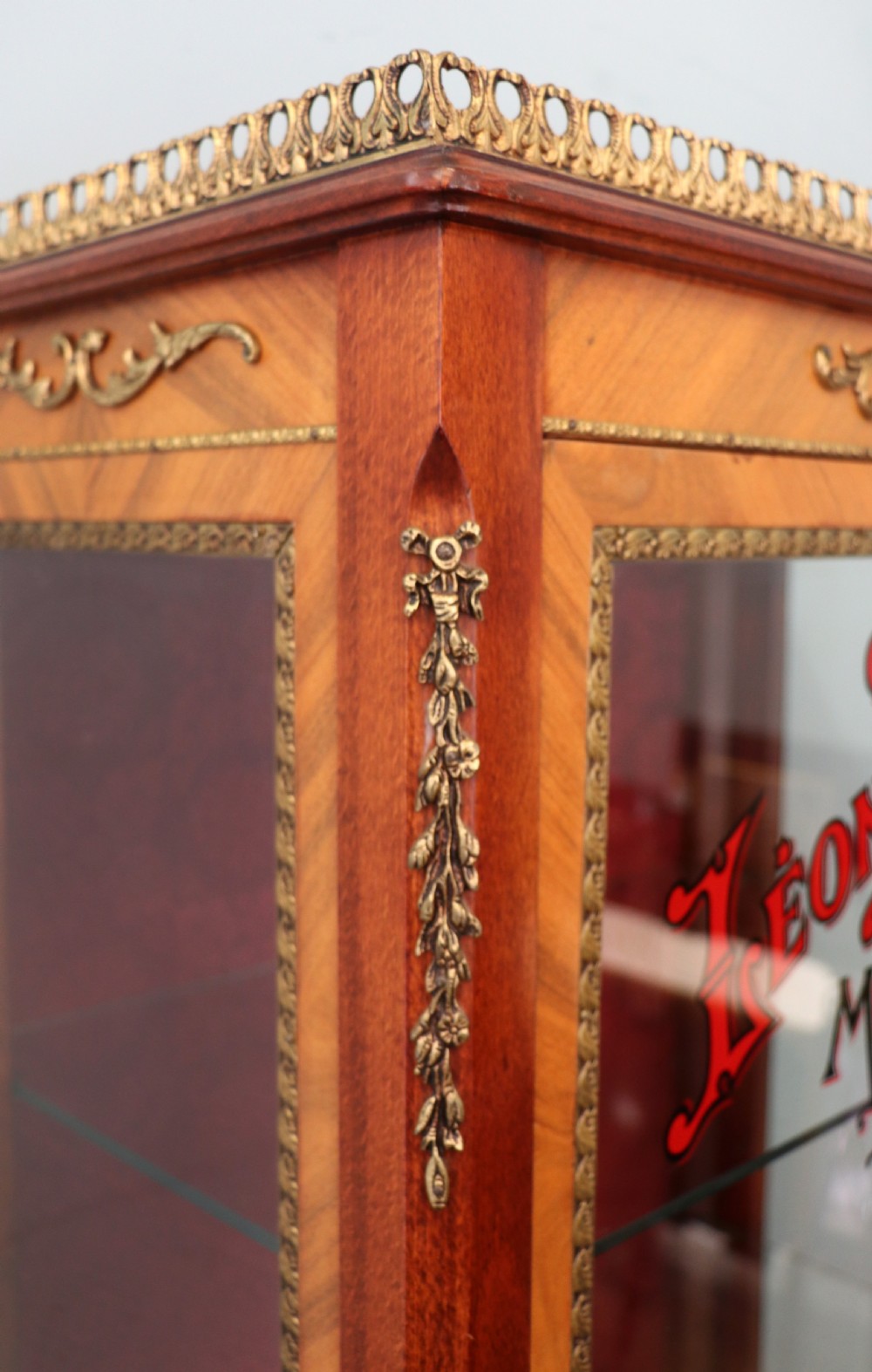 A French Vernis Martin style mahogany vitrine cabinet featuring a brass top gallery, glass shelves, red fabric lining, and a glazed door with gilded lettering. View X