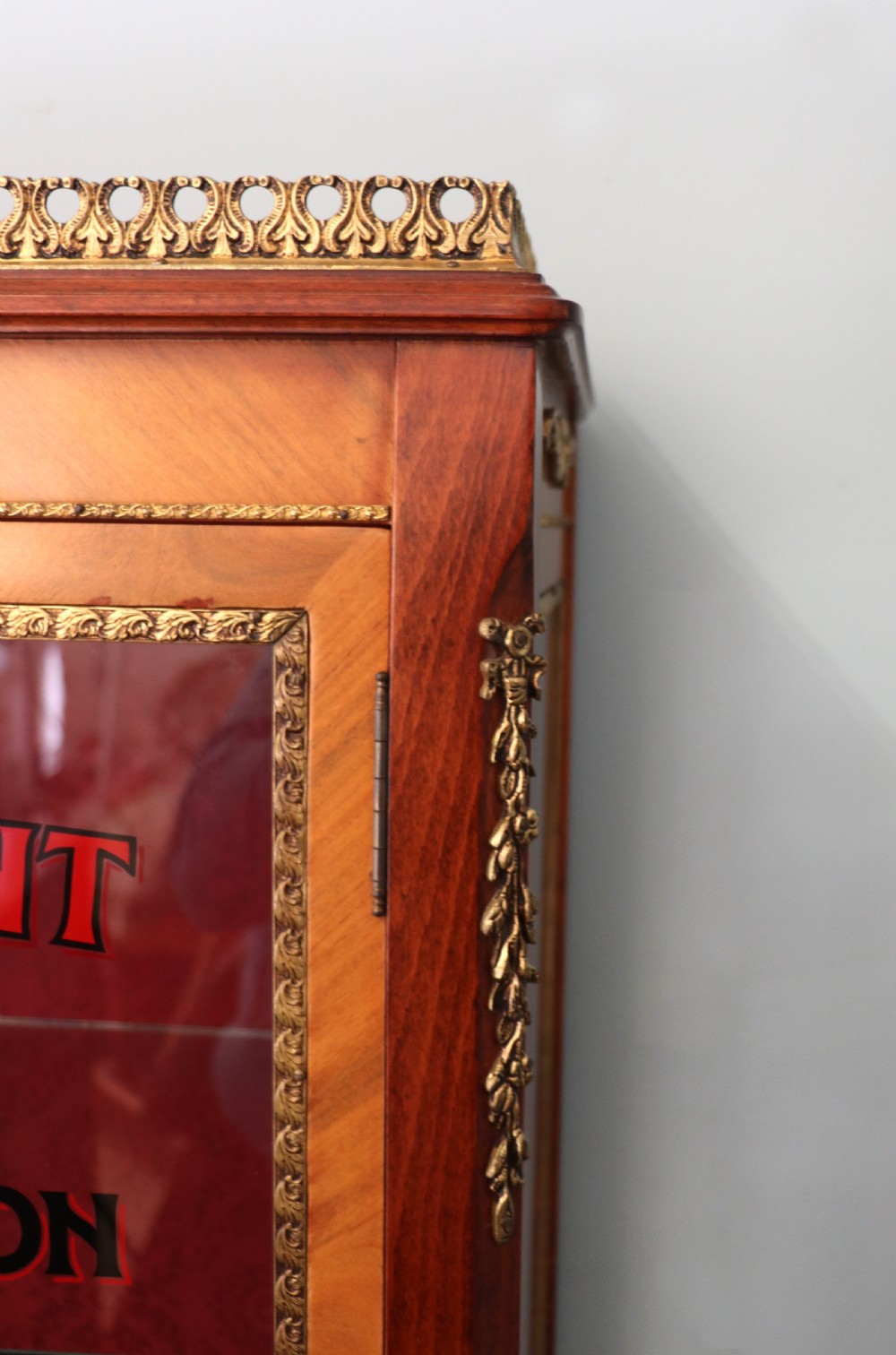 A French Vernis Martin style mahogany vitrine cabinet featuring a brass top gallery, glass shelves, red fabric lining, and a glazed door with gilded lettering. View XII