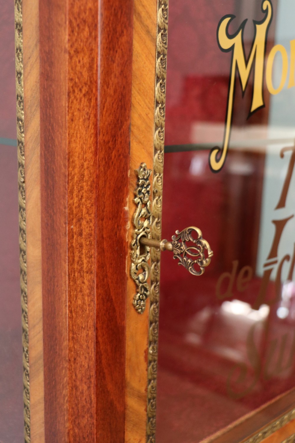 A French Vernis Martin style mahogany vitrine cabinet featuring a brass top gallery, glass shelves, red fabric lining, and a glazed door with gilded lettering. View XXV