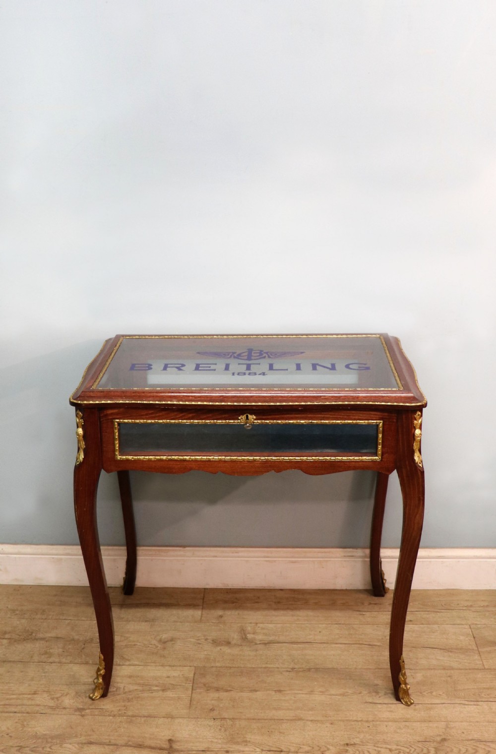 A French Louis XV style glazed bijouterie table in dark hardwood with cabriole legs, gilt metal mounts, and blue Breitling lettering on the glass top. View II