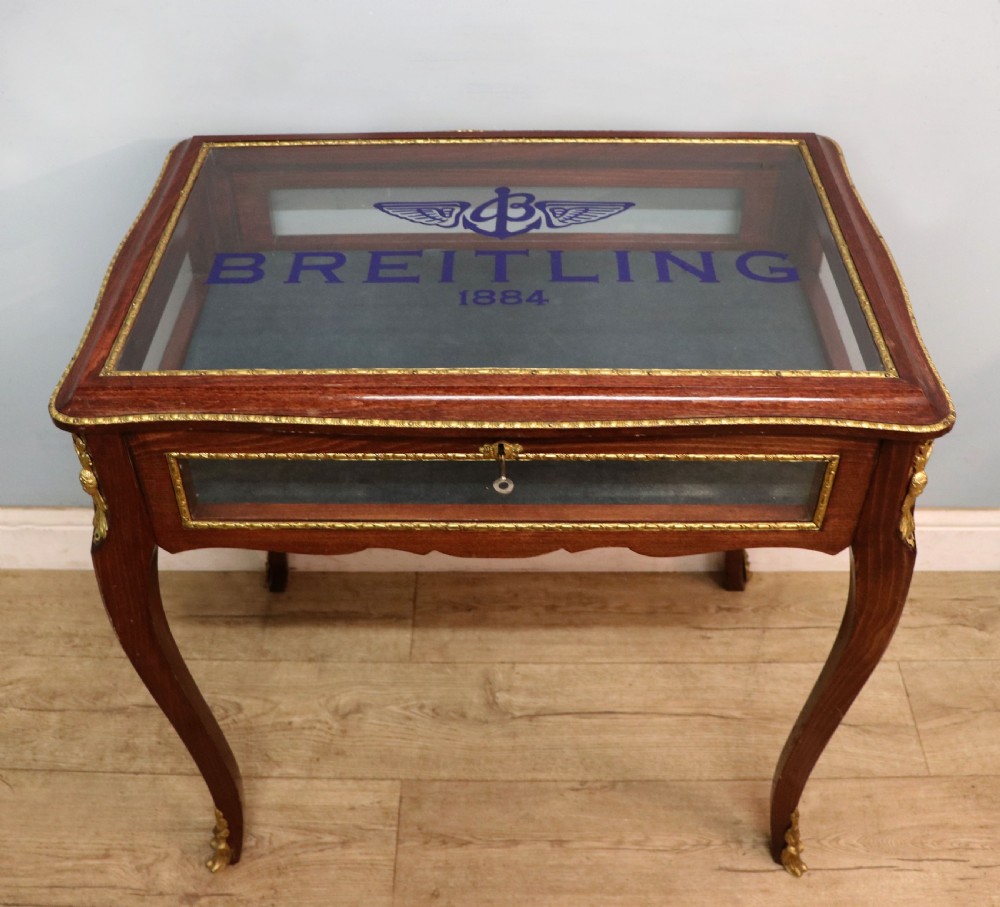 A French Louis XV style glazed bijouterie table in dark hardwood with cabriole legs, gilt metal mounts, and blue Breitling lettering on the glass top.