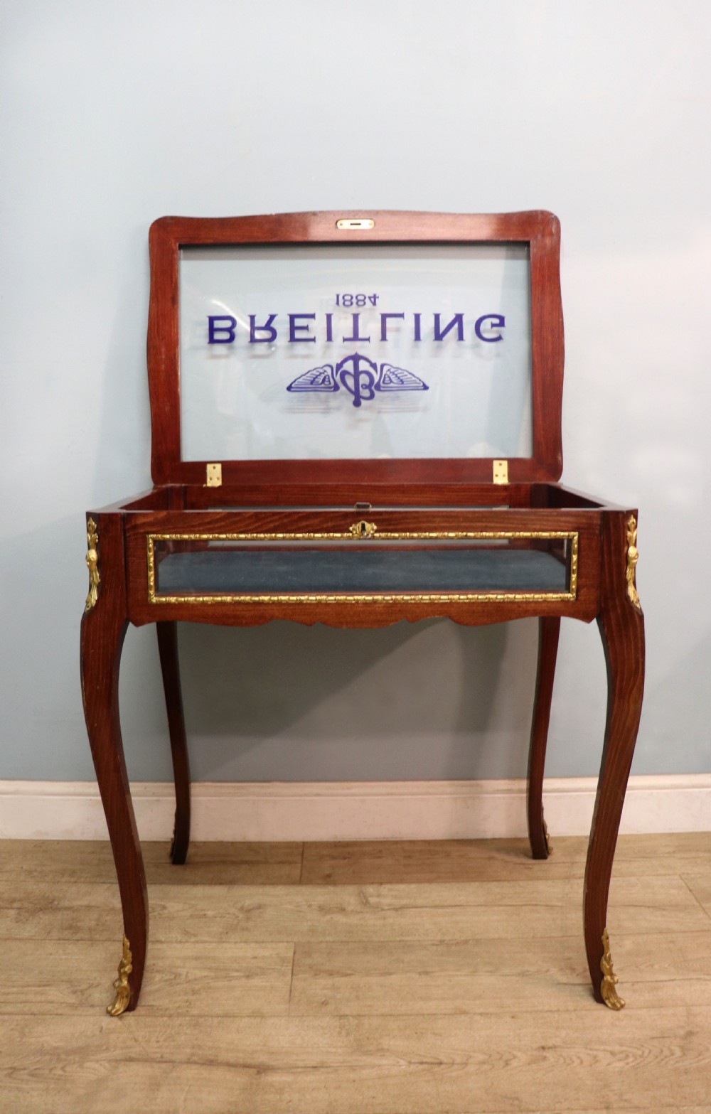 A French Louis XV style glazed bijouterie table in dark hardwood with cabriole legs, gilt metal mounts, and blue Breitling lettering on the glass top. View IV