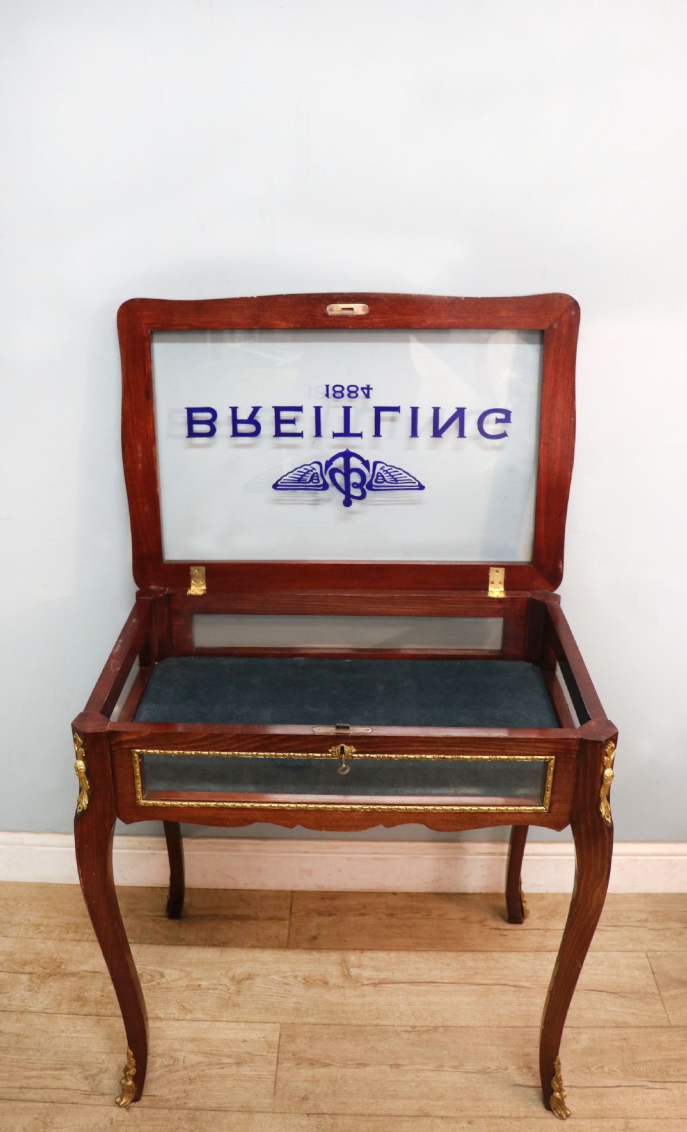 A French Louis XV style glazed bijouterie table in dark hardwood with cabriole legs, gilt metal mounts, and blue Breitling lettering on the glass top. View V