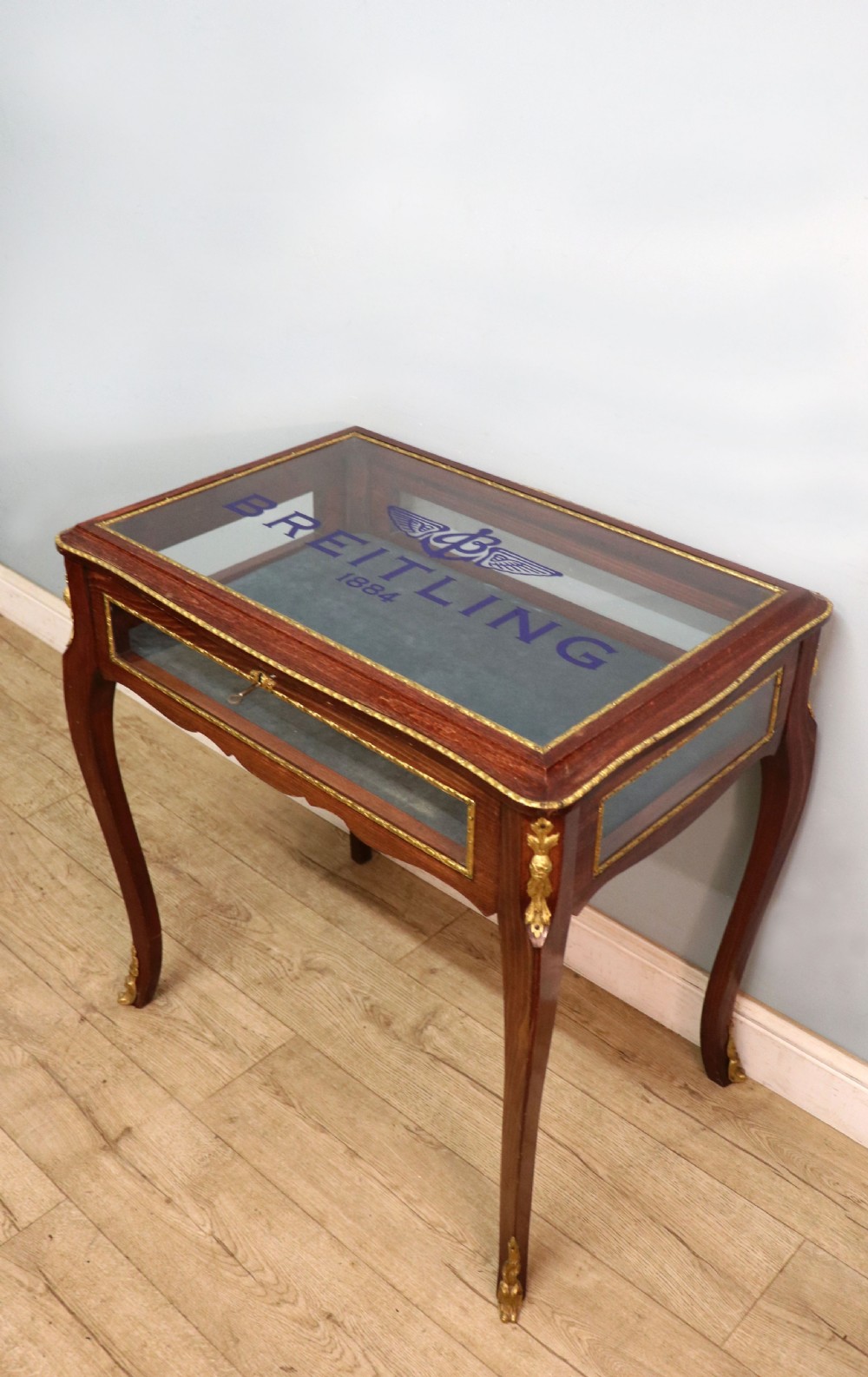 A French Louis XV style glazed bijouterie table in dark hardwood with cabriole legs, gilt metal mounts, and blue Breitling lettering on the glass top. View VII