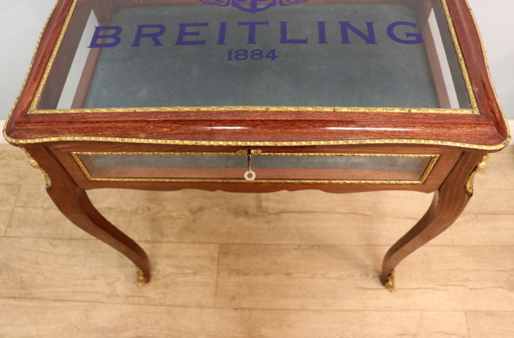 A French Louis XV style glazed bijouterie table in dark hardwood with cabriole legs, gilt metal mounts, and blue Breitling lettering on the glass top. View X
