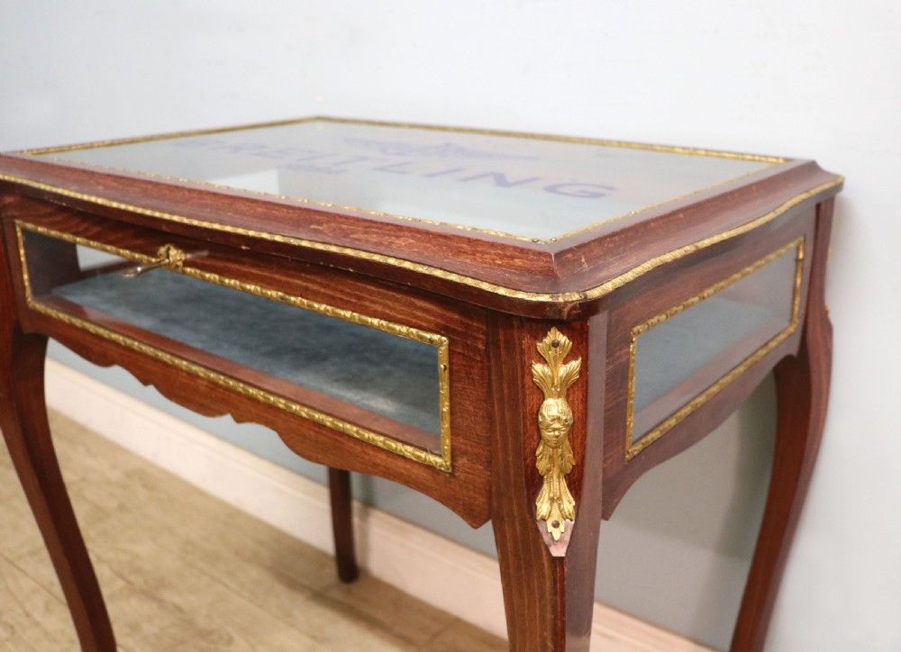 A French Louis XV style glazed bijouterie table in dark hardwood with cabriole legs, gilt metal mounts, and blue Breitling lettering on the glass top. View XII
